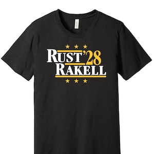 May include: Black t-shirt with a white graphic that says "Rust'28 Rakell" with gold stars.