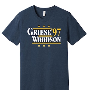 May include: Navy blue t-shirt featuring the text "Griese '97 Woodson" in white and yellow. The text is centered on the front, with three yellow stars above and below. The shirt has a crew neck and short sleeves, a classic design.