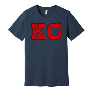 Può includere: Una t-shirt blu navy con una grafica rossa invecchiata delle lettere "KC" in un font universitario.