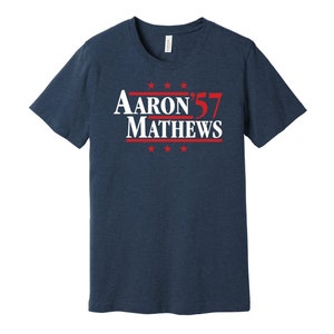 May include: Navy blue t-shirt with the text "Aaron Mathews 57" in white and red. The text is centered and includes red stars above and below the name. The shirt is made of a soft material.