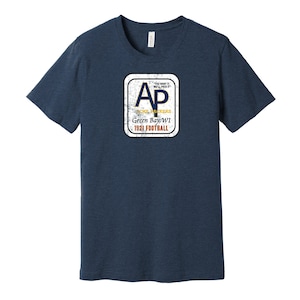 Puede incluir: Una camiseta azul marino con un logotipo de estilo vintage que presenta las letras "AP" en dorado y azul, con el texto "Green Bay WI 1921 FOOTBALL" debajo. El logotipo está rodeado por un borde blanco con un aspecto desgastado.