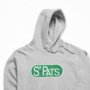 May include: A grey hooded sweatshirt with a front pocket and a drawstring hood. The sweatshirt displays the text "ST PATS" in green letters within a white and green oval. A casual style.