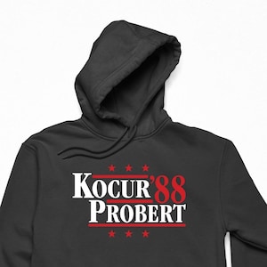 May include: Black hooded sweatshirt featuring the text "KOCUR 88 PROBERT" in white and red. The design includes red stars above and below the text. The sweatshirt has a front pocket and a drawstring hood.