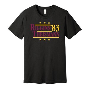 May include: Black t-shirt with a burgundy and gold graphic that reads "Riggins '83 Theisman" with four gold stars above and below the text.