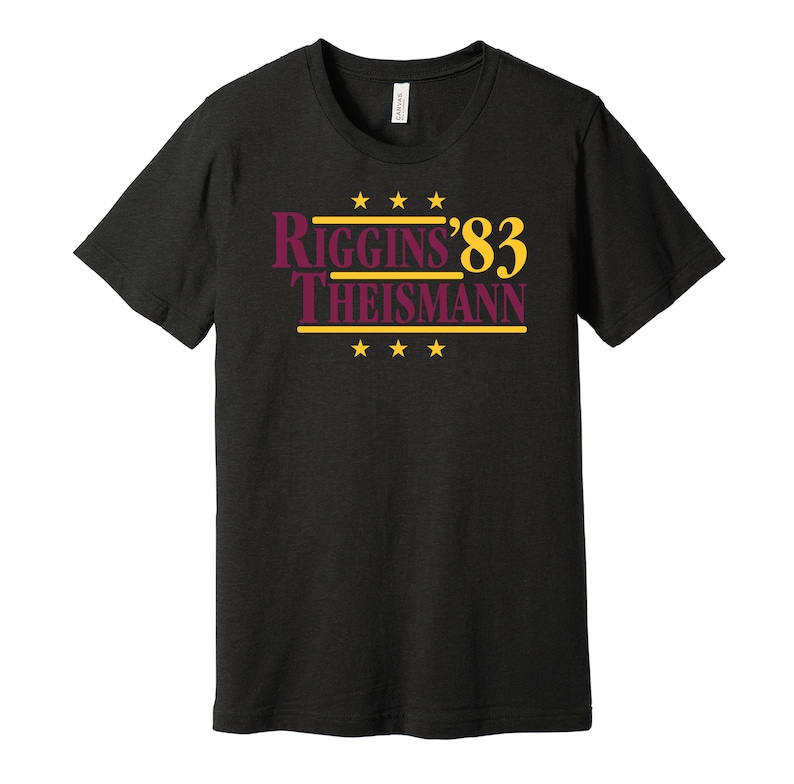 May include: Black t-shirt with a burgundy and gold graphic that reads "Riggins '83 Theisman" with four gold stars above and below the text.