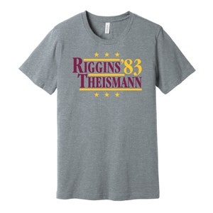 May include: A heather gray t-shirt with a burgundy and gold graphic that reads "Riggins '83 Theisman" with five gold stars above and below the text.
