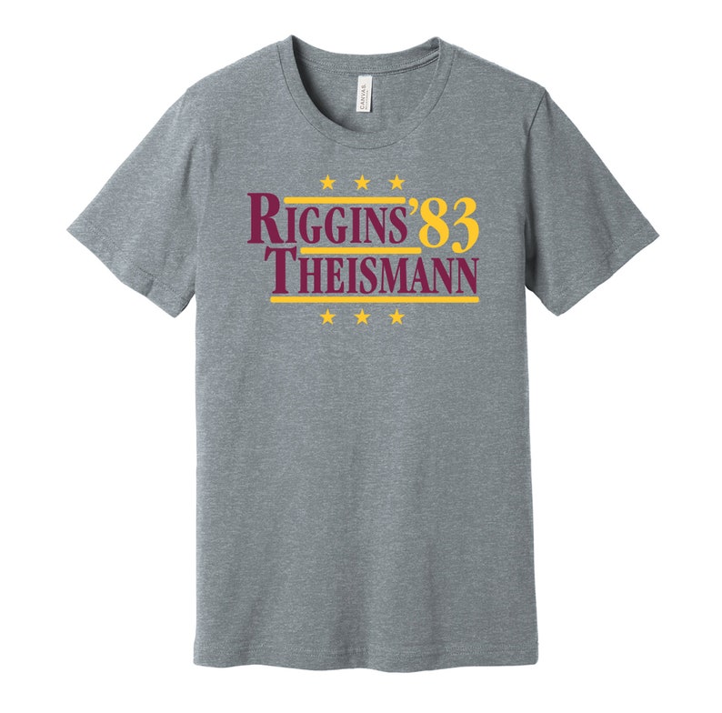 May include: A heather gray t-shirt with a burgundy and gold graphic that reads "Riggins '83 Theisman" with five gold stars above and below the text.