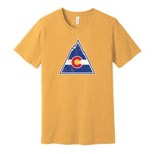 May include: A yellow t-shirt with a distressed graphic of a mountain range and the Colorado state flag. The flag is blue, white, and red with a large "C" in the center.
