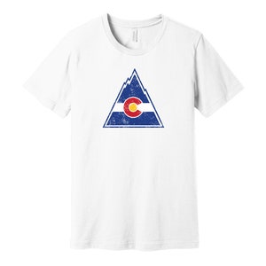 May include: White t-shirt with a distressed graphic of a mountain range with the Colorado flag inside. The flag is red, yellow, and blue with a large "C" in the center.