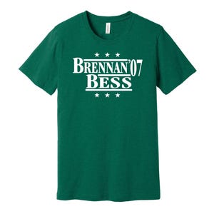 May include: A green t-shirt featuring the white text "Brennan '07 Bess", with three stars above and below. The shirt has a crew neck and short sleeves.