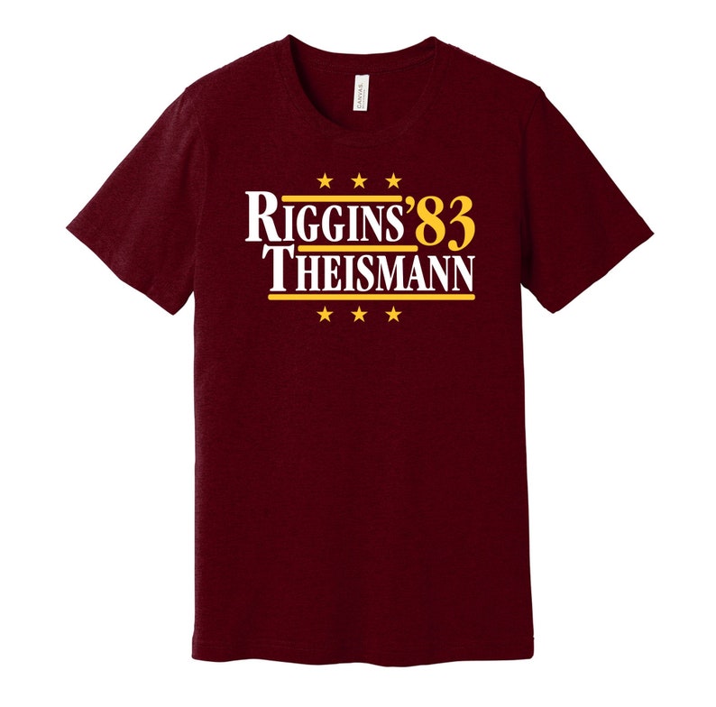 May include: Burgundy t-shirt with a white graphic that says "Riggins'83 Theisman" in gold lettering with five gold stars above and below the text.