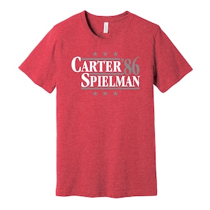 May include: Red t-shirt featuring the text "Carter '86 Spielman" in white and grey. The design includes stars above and below the text. The shirt is made of a soft, textured material.