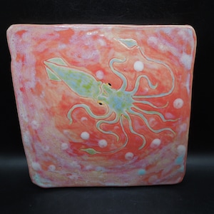 May include: Square ceramic dish with a hand-painted design of a squid. The squid is light blue and green, with yellow eyes, set against a coral-colored background with white bubbles. The dish has a slightly irregular shape.