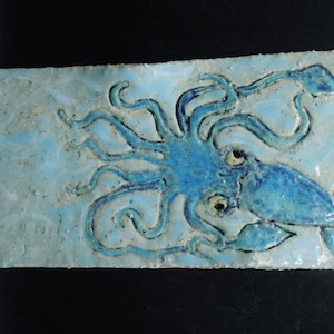 May include: A blue ceramic tile with a raised image of a squid. The squid has a light blue body and yellow eyes. The tile has a light blue background.