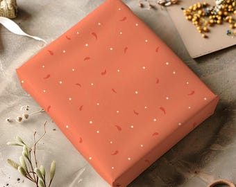 May include: A square gift wrapped in orange paper with a pattern of white dots and red crescent shapes. Other wrapped gifts, flowers, and craft supplies are arranged around it on a gray surface.