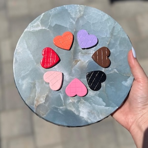 May include: A circular arrangement of heart-shaped objects in various colours, including red, orange, purple, brown, black, pink, and light pink, on a blue-green stone surface. Each heart has a textured pattern. The stone is round.