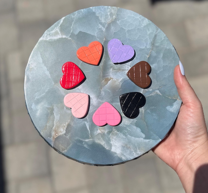 May include: A circular arrangement of heart-shaped objects in various colours, including red, orange, purple, brown, black, pink, and light pink, on a blue-green stone surface. Each heart has a textured pattern. The stone is round.