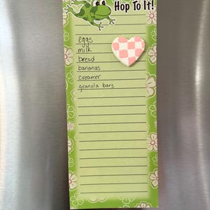 May include: A green notepad with a frog illustration and the text "Hop To It!". The notepad has a list of grocery items and a pink and white checkered heart-shaped magnet. Floral designs decorate the edges.