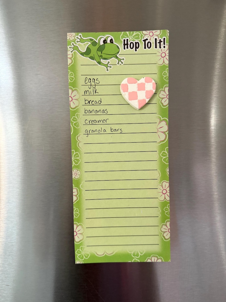May include: A green notepad with a frog illustration and the text "Hop To It!". The notepad has a list of grocery items and a pink and white checkered heart-shaped magnet. Floral designs decorate the edges.