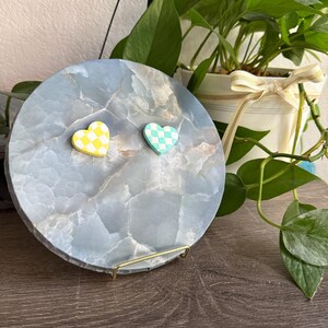 May include: A circular, pale blue decorative tray with a marbled effect. Two heart-shaped magnets, one yellow and white chequered, the other teal and white chequered, are attached. The tray is displayed on a gold metal stand.