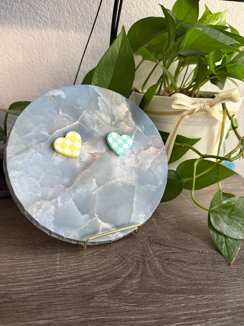 May include: A circular, pale blue decorative tray with a marbled effect. Two heart-shaped magnets, one yellow and white chequered, the other teal and white chequered, are attached. The tray is displayed on a gold metal stand.