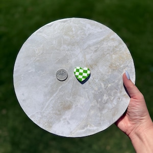 May include: A round, grey marble-effect serving tray with a green and white chequered heart-shaped object and a coin. The tray is held by a person's hand against a blurred green background.