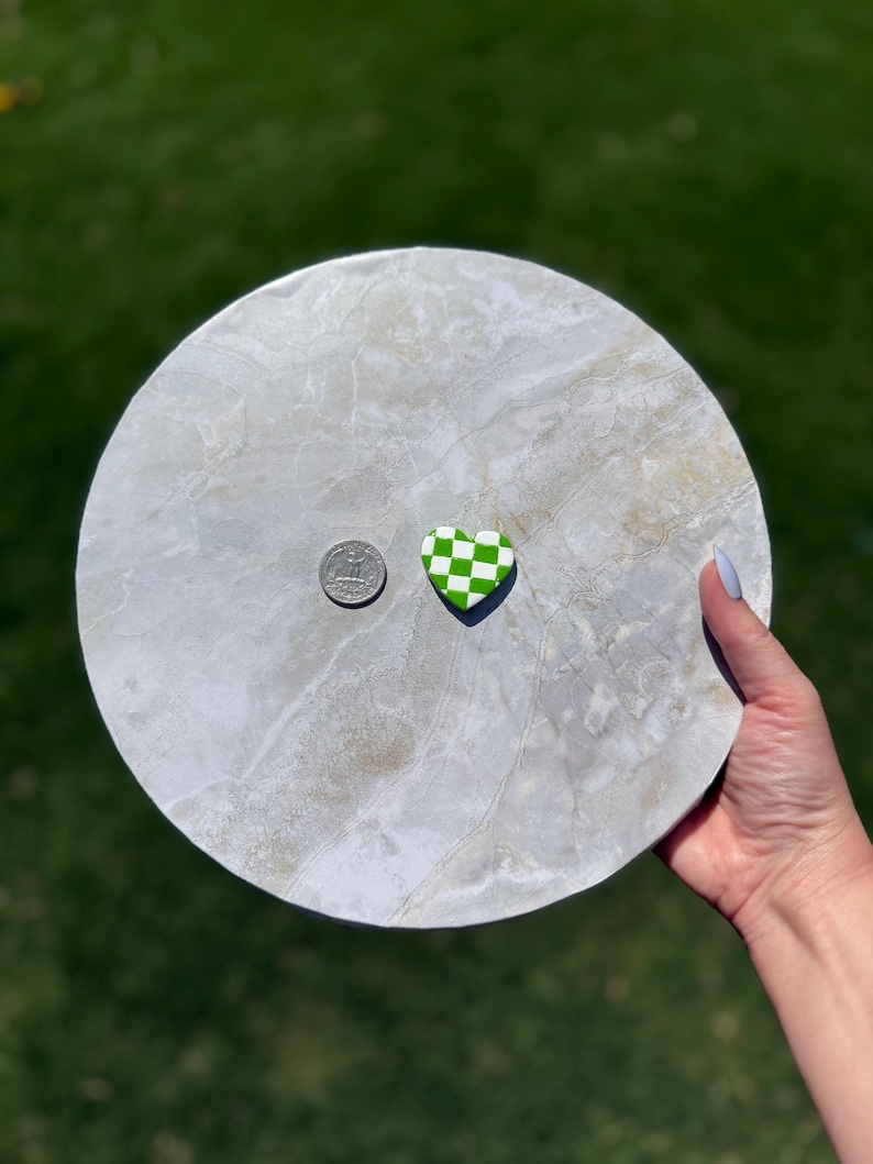 May include: A round, grey marble-effect serving tray with a green and white chequered heart-shaped object and a coin. The tray is held by a person's hand against a blurred green background.