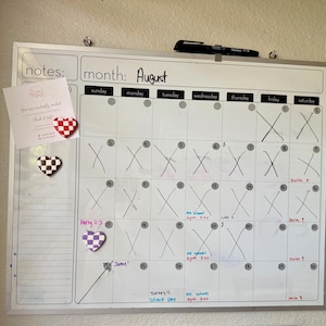 May include: A white magnetic calendar board showing the month of August. The calendar has handwritten notes and crossed-out dates. Heart-shaped magnets in red and white, black and white, and purple and white are attached to the board.