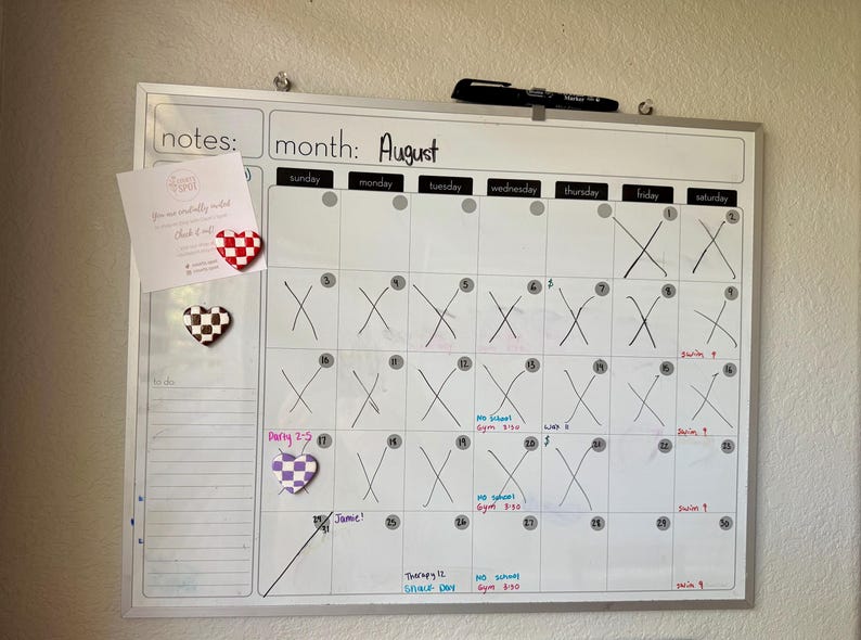 May include: A white magnetic calendar board showing the month of August. The calendar has handwritten notes and crossed-out dates. Heart-shaped magnets in red and white, black and white, and purple and white are attached to the board.