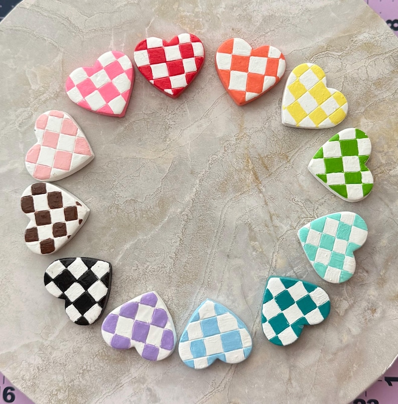 May include: A circular arrangement of heart-shaped decorations. Each heart is decorated with a checkerboard pattern in a range of colours: pink, red, orange, yellow, green, teal, blue, purple, brown, and black. The hearts are displayed on a neutral surface.
