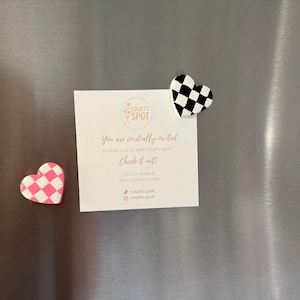 May include: Two heart-shaped magnets with chequered patterns, one pink and white, the other black and white, are attached to a metallic surface. A white card with text and a logo is also attached, inviting viewers to shop on Etsy.