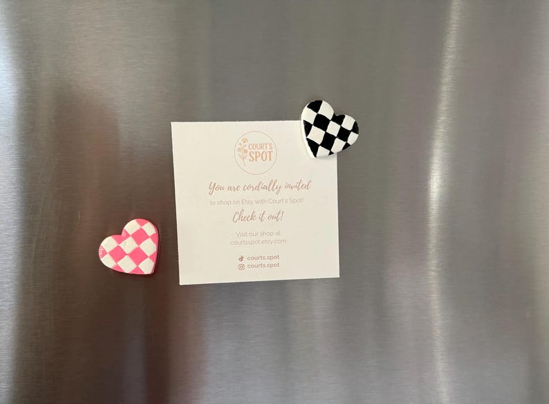 May include: Two heart-shaped magnets with chequered patterns, one pink and white, the other black and white, are attached to a metallic surface. A white card with text and a logo is also attached, inviting viewers to shop on Etsy.