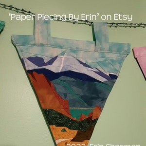 May include: A triangular fabric wall hanging with a mountain scene. The fabric is pieced together in different colors and patterns, including blue, green, brown, and white. The hanging has a loop at the top for hanging. The text "2022 Erin Sherman" is printed at the bottom of the hanging.