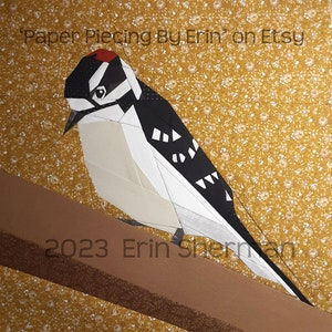 May include: A paper pieced pattern of a black and white woodpecker with a red crown perched on a brown branch. The background is a yellow floral print fabric. The text "2023 Erin Sherman" is visible in the lower left corner.