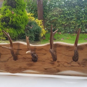 May include: A wooden coat rack with five antler hooks, mounted on a wall. The rack is made from light-coloured, weathered wood with a natural shape. The antlers are a dark brown colour.