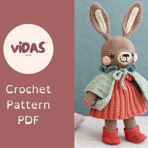 Op de afbeelding: Een gehaakte konijnenpop met lange oren, gekleed in een koraalkleurige jurk, een lichtgroene cape en rode laarzen. De pop heeft een bruine kop en lichaam met roze wangen. De afbeelding bevat de tekst "VIDAS Studio" en "Crochet Pattern PDF".