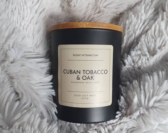 Może przedstawiać: Czarny słoik na świecę z drewnianą pokrywką. Etykieta zawiera napis "Scent of Sanctum", "Cuban Tobacco & Oak", "Handmade with Care", "100% Soy Wax" i "220g".