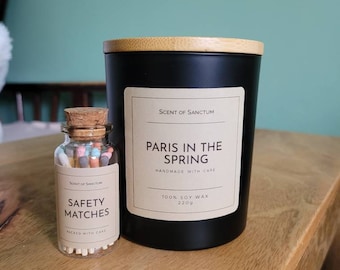 May include: A black candle with a wooden lid and a label that reads "Scent of Sanctum Paris in the Spring Handmade with Care 100% Soy Wax 220g". A small glass jar with a cork lid and a label that reads "Scent of Sanctum Safety Matches Packed with Care" is next to the candle.