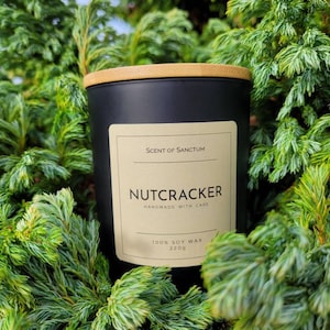 Può includere: Una candela nera con un coperchio in legno e un'etichetta che recita "Scent of Sanctum" e "Nutcracker Handmade with Care 100% Soy Wax 220g". La candela è circondata da fogliame verde.