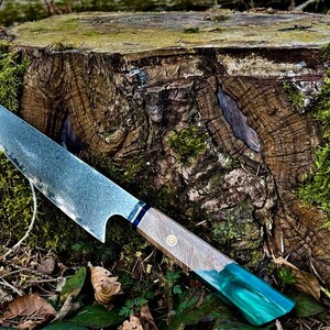 Könnte beinhalten: Ein handgemachtes Messer mit blauem und grünem Griff, das auf einem mit Moos bewachsenen Baumstumpf liegt.