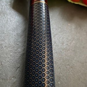 May include: A close-up of a black and blue patterned knife handle with a red and gold accent. The handle is made of a textured material and has a metallic finish.