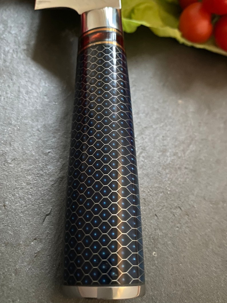 May include: A close-up of a black and blue patterned knife handle with a red and gold accent. The handle is made of a textured material and has a metallic finish.