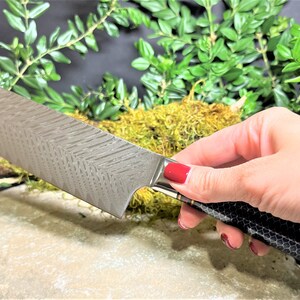 May include: A silver chef's knife with a patterned blade and a black and gold handle. The knife is resting on a rock with green foliage in the background.