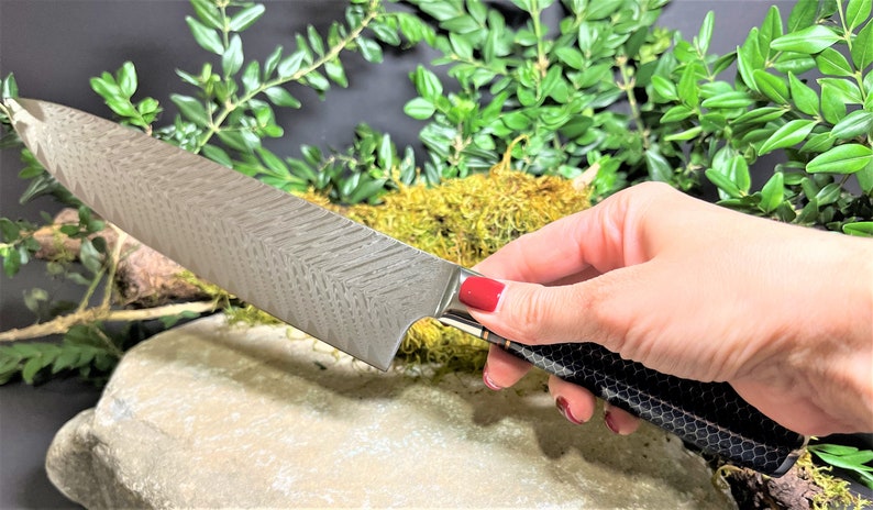 May include: A silver chef's knife with a patterned blade and a black and gold handle. The knife is resting on a rock with green foliage in the background.