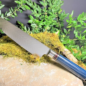 May include: A silver chef's knife with a blue and gold patterned handle. The knife is resting on a rock with moss and green foliage.