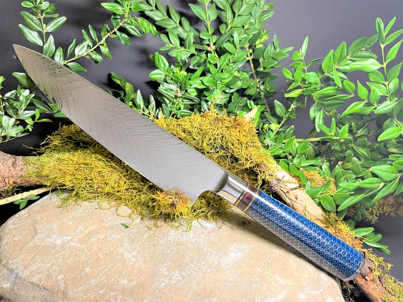May include: A silver chef's knife with a blue and gold patterned handle. The knife is resting on a rock with moss and green foliage.