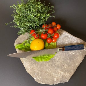 May include: A close-up of a gray stone slab with a lemon, cherry tomatoes, and a sprig of greenery. A silver knife with a blue handle is resting on the stone.