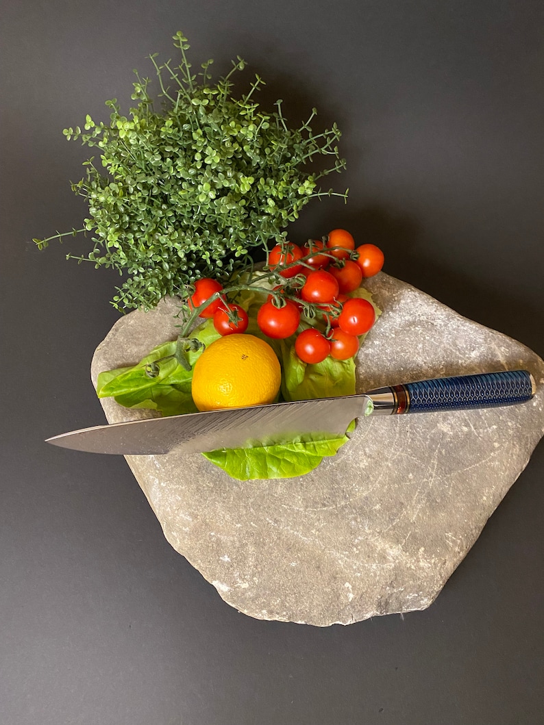 May include: A close-up of a gray stone slab with a lemon, cherry tomatoes, and a sprig of greenery. A silver knife with a blue handle is resting on the stone.