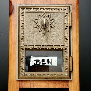 May include: A small wooden mailbox with a bronze-colored metal door. The door features a keyhole and a decorative starburst design. The name "BEN" is written on a small white card visible through a window.