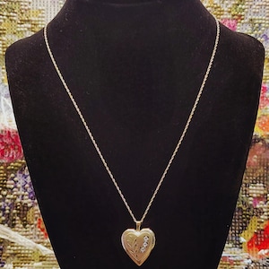 May include: A gold-toned heart-shaped locket pendant suspended on a thin gold-toned rope chain. The locket features an engraved floral design on the left side and three small, clear, sparkling stones set vertically on the right side of the heart.